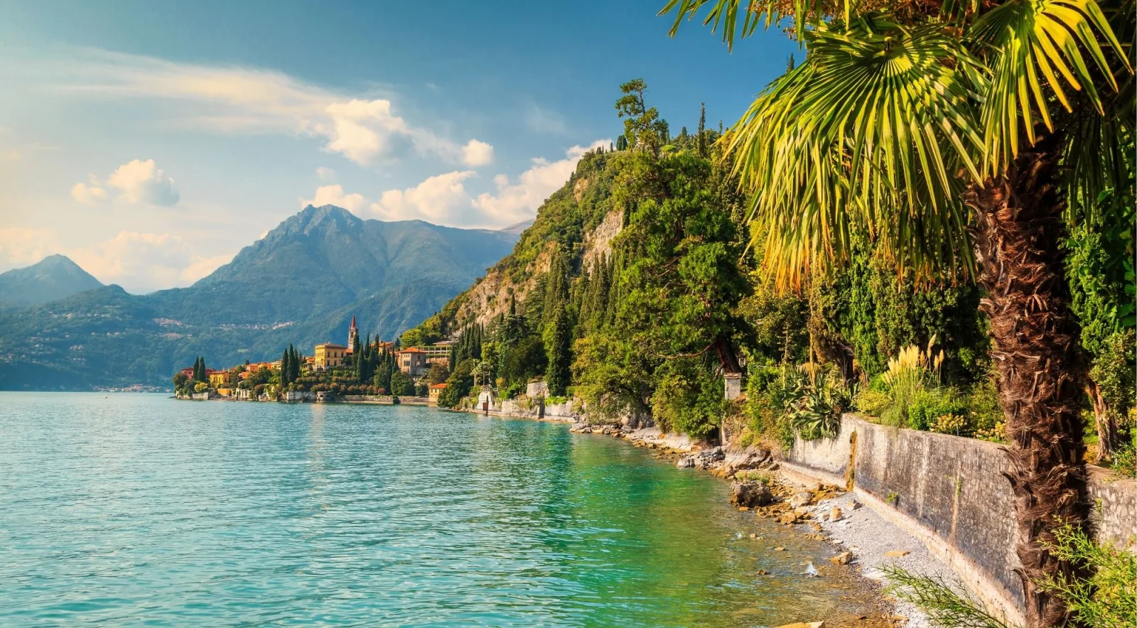The Villa Monastero lakefront walk under palms, with Varenna's bell tower on the headland beyond