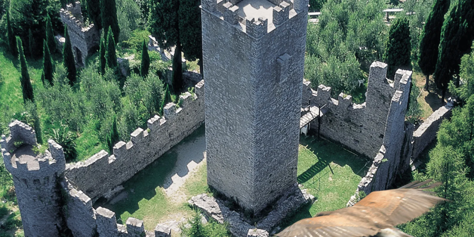Castello di Vezio from the air — the square keep inside its curtain wall, a hawk crossing the frame